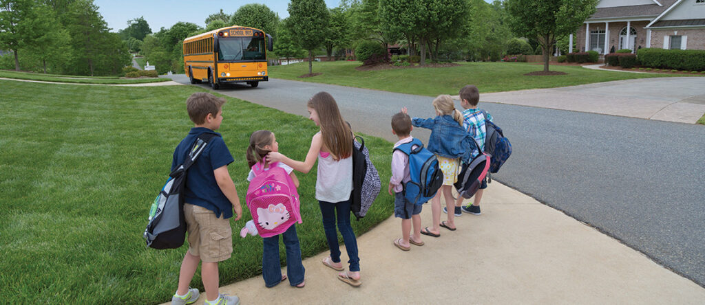 Kids standing on sidewalk with green grass in front and black pavement to the right. A yellow school buses approaches on the pavement