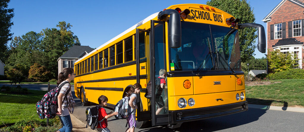 Children entering yellow school bus in a residential community with a brick house on the right and green trees and grass in the background