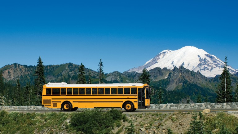 Yellow school bus on a road with blue skies, green trees, and a white mountain in the background covered in snow