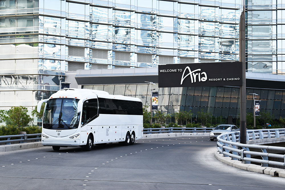 White charter bus driving on black pavement with large glass building in background.